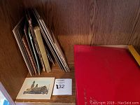 A collection of antique books, mounted pictures and photographs stacked on a wooden surface, featuring one image of a historic building and a red hardbound book.