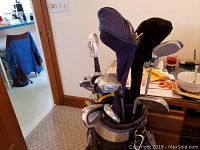 Photo showing golf clubs including irons, woods and putter in a silver and black golf bag inside a domestic room.
