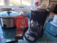 Photo showing Rival slow cooker with glass lid, a Mr. Coffee drip coffee maker with glass carafe, and two packets of coffee on counter.