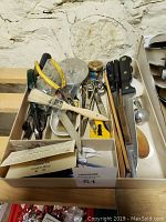 Photo showing a variety of scissors, kitchen knives, pliers, a paintbrush, small tools and utensils arranged in cardboard boxes and a drawer divider against a white textured wall.