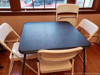 Bridge table with four white folding chairs set around it near window
