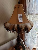 Front view of the tan fabric lamp shade decorated with a trim of multi-toned brown and white feathers, showing the overall shape and feather details.