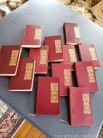 12 classic books laid out on a table, showing red leather covers with a gold gilt decorative design.