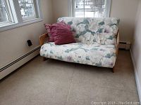Wood frame futon with floral mattress and red and purple pillows, placed in a corner with windows.