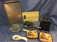 Photo of metal baking tray, wooden cutting board, stainless steel egg poacher pan, mesh strainer, black Hamilton Beach toaster, white ceramic spoon rest with fruit design, and two ceramic pot holders with fruit design