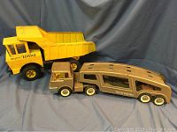 Wide view showing both the yellow Mighty Tonka dump truck and the beige Structo Auto Haul car carrier trailer with cab, placed on grey cloth backdrop.