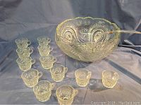 Vintage pressed glass punch bowl with 12 matching cups arranged on a surface and ladle resting inside the bowl. Clear glass with intricate starburst and geometric pressed patterns.