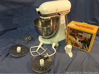 KitchenAid stand mixer with mixing bowl, wire whisk, dough hook, flat beater, slicing and shredding discs, and food grinder attachment box.