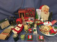 Wide view showing various Christmas decorations including vintage Santa face, green glitter trees, red glass candle holders, stockings, and assorted tins and boxes spread on blue backdrop.