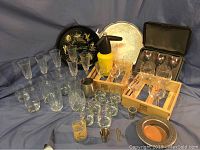 Wide view showing multiple glassware items, vintage yellow seltzer bottle, black Lake George souvenir tray, and silver serving tray on blue fabric background.