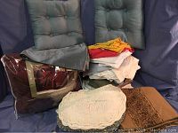 Lot of linens displayed against a blue backdrop, including two tufted blue-green throw pillows, a burgundy comforter set in packaging, and a stack of folded sheets with lace doilies and placemats in front.