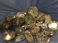 Photo showing a broad assortment of silver plate trays, bowls, teapots, and covered dishes with intricate designs and patterns.