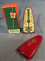 Photo showing Wittner Taktell vintage mechanical metronome, red plastic body with pendulum arm and tempo scale, original colorful cardboard box with Wittner logo, and red triangular plastic cover with logo.
