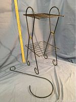 Metal plant stand with two wire shelves and curled legs shown with tape measure for size reference (height approx. 27.75 inches).
