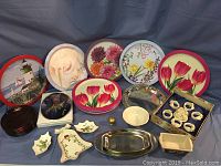 Photo showing an assortment of decorative round trays with floral, lighthouse, and shell patterns, red glass plates, and small home decor pieces including candle stick and napkin holders.