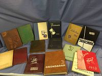 Overview of 14 vintage yearbooks from various schools with diverse cover designs and colors arranged on a gray fabric background.