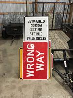 View showing stacked street signs including the white 'RESIDENTIAL STREETS POSTED UNLESS OTHERWISE' sign and red 'WRONG WAY' sign, metal signs resting on a wooden floor.