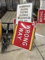 Five metal street signs stacked against chain link fencing indoors, includes wrong way, residential streets posted, and other signs.