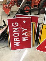 Photo showing red metal Wrong Way sign leaning against other signs and equipment.
