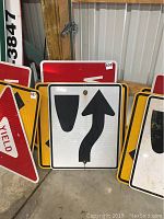 Photo showing several metal street signs including 'Yield' and lane direction signs with arrows and shield symbol