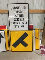 Photo showing various metal street signs stacked, including a large white Speed Limit 25 sign and yellow T-intersection sign.