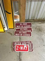 Seven metal signs, including three 'All Dogs Must Be Leashed', three 'Keep Off Ice No Ice Skating or Ice Fishing', and one '2-Way' sign stacked on a cement floor against a wooden post with visible wear and tape.