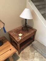 Wooden side table with lamp on top, showing wear on table surface, positioned near stairs.
