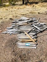 Pile of over 50 wooden stakes stacked outdoors on gravel with dry grass in background