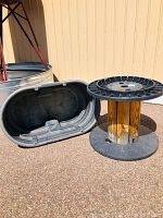 Photo showing large black Rubbermaid water trough and empty spool with wooden core and black plastic ends on outdoor ground against tan wall.