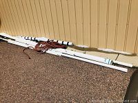 Seven white PVC pipes arranged on the ground outside along a yellow wall, with a coiled brown garden hose beside them.