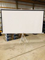 Full view of the white Elite Screens projector screen mounted on a white tripod stand in a warehouse setting.
