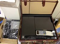 Overview photo showing vintage Bell South mobility phone integrated into a brown leather briefcase and AT&T wired phone box with plastic wrapped phone handset and buttons visible.