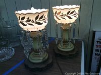 Pair of brass-bottom lamps with ceramic scalloped shades featuring black floral designs and hanging crystal prisms placed side by side on a table.