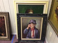 Image of two paintings leaning on wall: boy rubbing eye in black frame with cream border, girl holding fruit basket partially visible behind