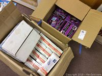 Two open boxes containing multiple boxed items: one box with purple boxes of binoculars labeled 4.5x40mm, and another box with white and red labeled Turbo Steamer irons boxes along with a small white box likely containing moist towelettes.