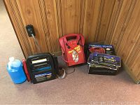 Full view of all items including Power Pack 12V power source in red, black Professional PowerStation PS1100 jump starter with clamps attached, blue gallon bottle of windshield washer fluid, and license plate frames with fasteners.