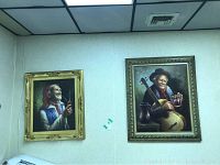 Two framed oil paintings on wall, showing elderly male portraits, one smoking a pipe and the other holding guitar and mug.