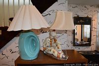 View of both lamps on a wooden surface under stairs, showing the turquoise ceramic lamp with pleated white shade and the figurative lamp with a seated figure in colorful clothing and a bell-shaped fabric shade.
