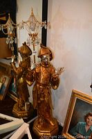 Full view of the 71-inch tall gold toned Chinese figure in traditional dress holding a prism-adorned staff, positioned among framed paintings and lamps.
