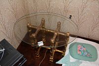 Oval glass top end table with gold-toned metal base in faux bamboo design on brown carpet and floral wallpaper background
