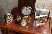 Seven desk clocks of various styles on a wooden table near window, includes brass, glass, and wood parts
