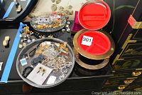 Two round metal jewelry boxes side by side on black surface, one with red velvet interior and brass rim