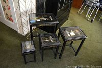 Full set of four black lacquer nesting tables with beveled glass tops showing Asian themed raised relief figures and gold accent decoration.