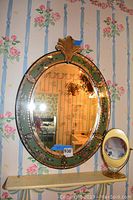 Full front view of the large hand painted oval wall mirror with floral decoration and brass details, visible crack and missing piece at top, and the small brass side vanity mirror to the right.