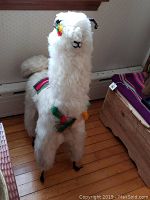 Front view of soft llama figurine with colorful yarn tassels and woven saddle blanket on wooden floor.