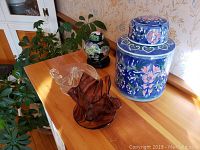 Photo showing clear glass juicer, brown glass bowl/vase, large blue floral ceramic jar, and smaller black floral ceramic jar on wooden stand on wooden surface with plant in background.