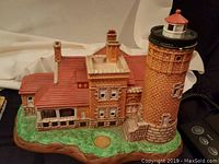 Close-up of a large lighthouse sculpture with an attached building featuring multiple chimneys and windows on a green grass-like base, cable attached for lighting.