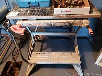 Black and Decker Workmate 426 portable workbench with two orange clamp handles, metal frame, footrest platform, and work surface marked by paint and use.