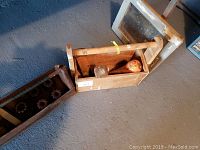 Overview photo showing an antique wooden carpenter's caddy containing a wooden mallet, round wooden weight, and part of a wooden framed gear set next to it on the floor.
