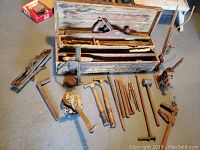 Overview photo of the antique wooden tool box open showing two hand planes inside and many tools arranged in front including hammers, braces, chisels, clamps, and wrenches on a concrete floor.